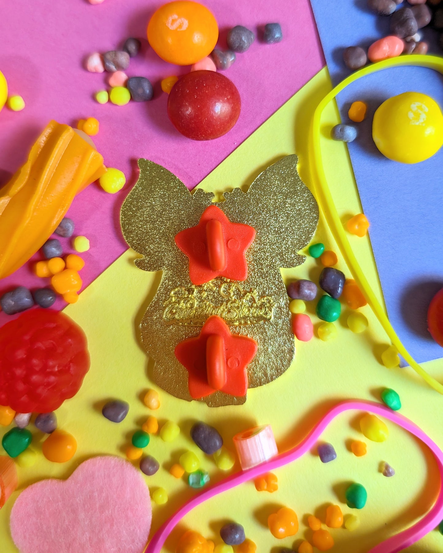Back of enamel pin featuring red rubber clasps shaped like stars set against a colourful background with rainbow candies