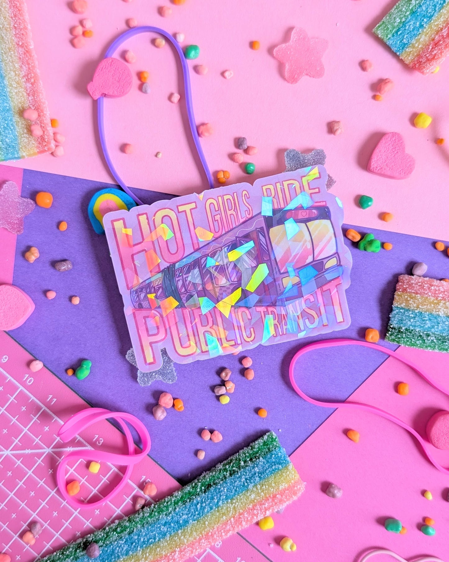 Colorful sticker on pink and purple background says "Hot Girls Ride Public Transit" with illustration of transit bus. Surrounded by rainbow candies and shapes.