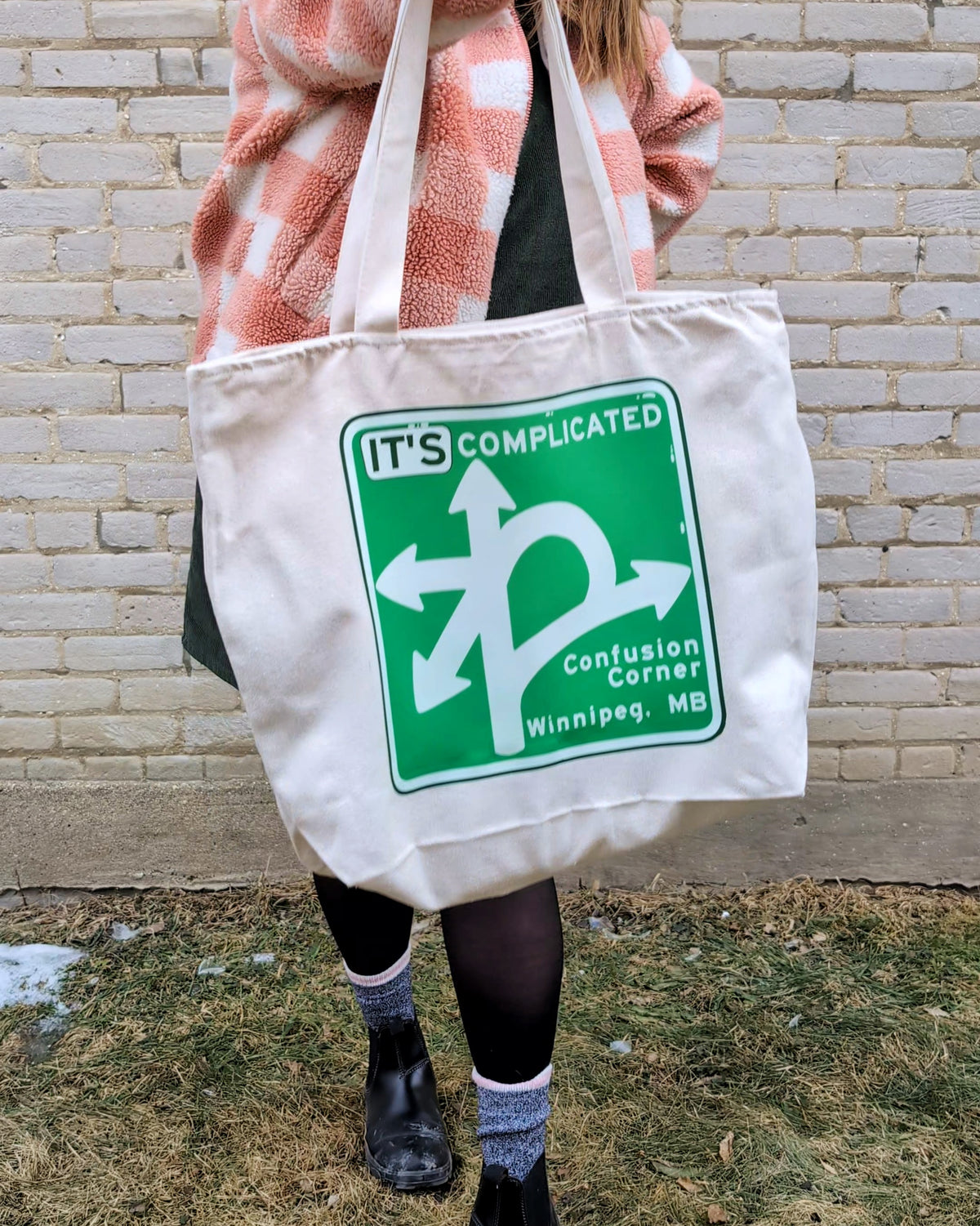 White canvas tote bag printed with a Confusion Corner road sign saying it's complicated is being held up by a woman
