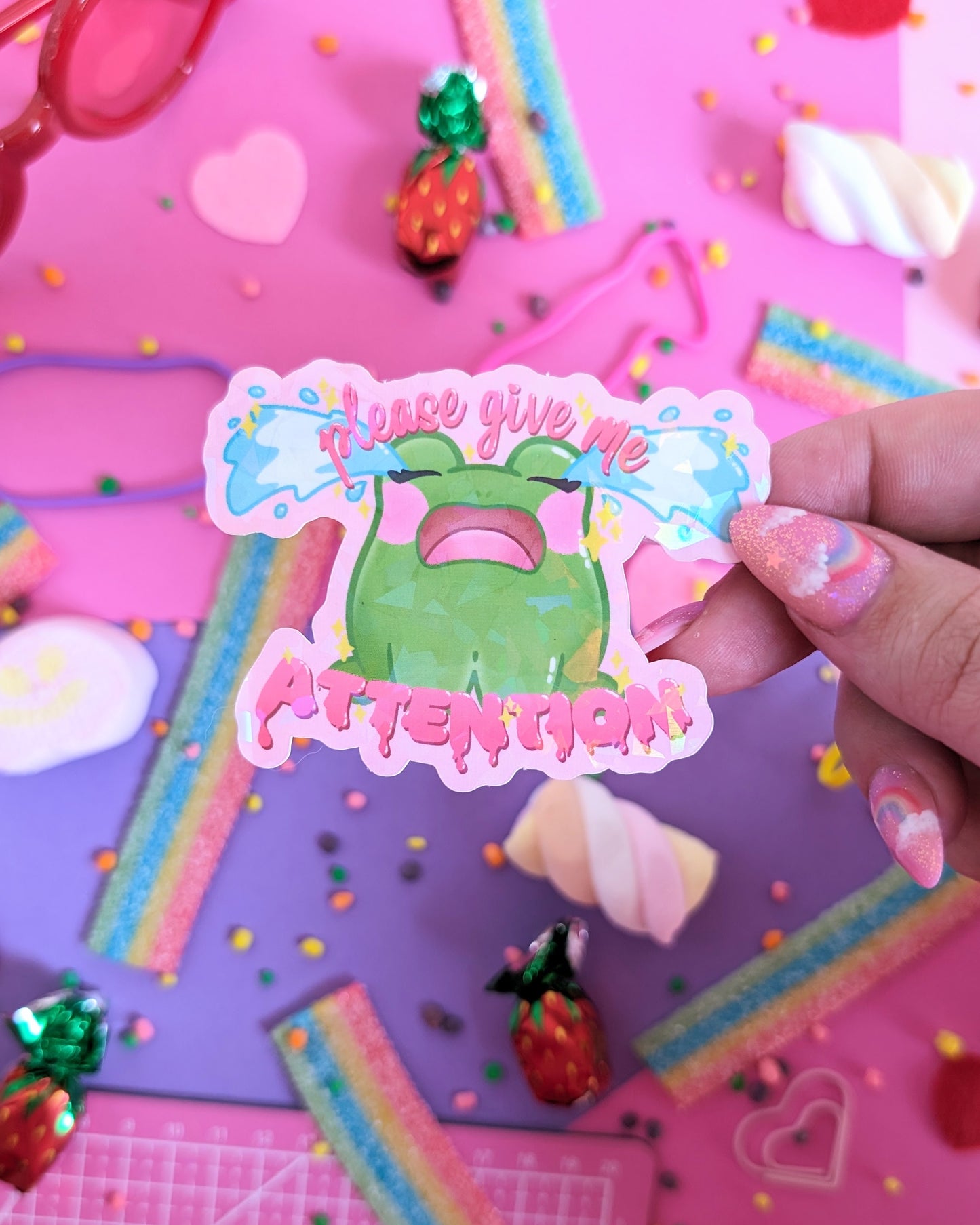 Hand with pink nails holding a colorful sticker with a green cartoon frog and the text "please give me ATTENTION" on a vibrant pink and purple candy-themed background.