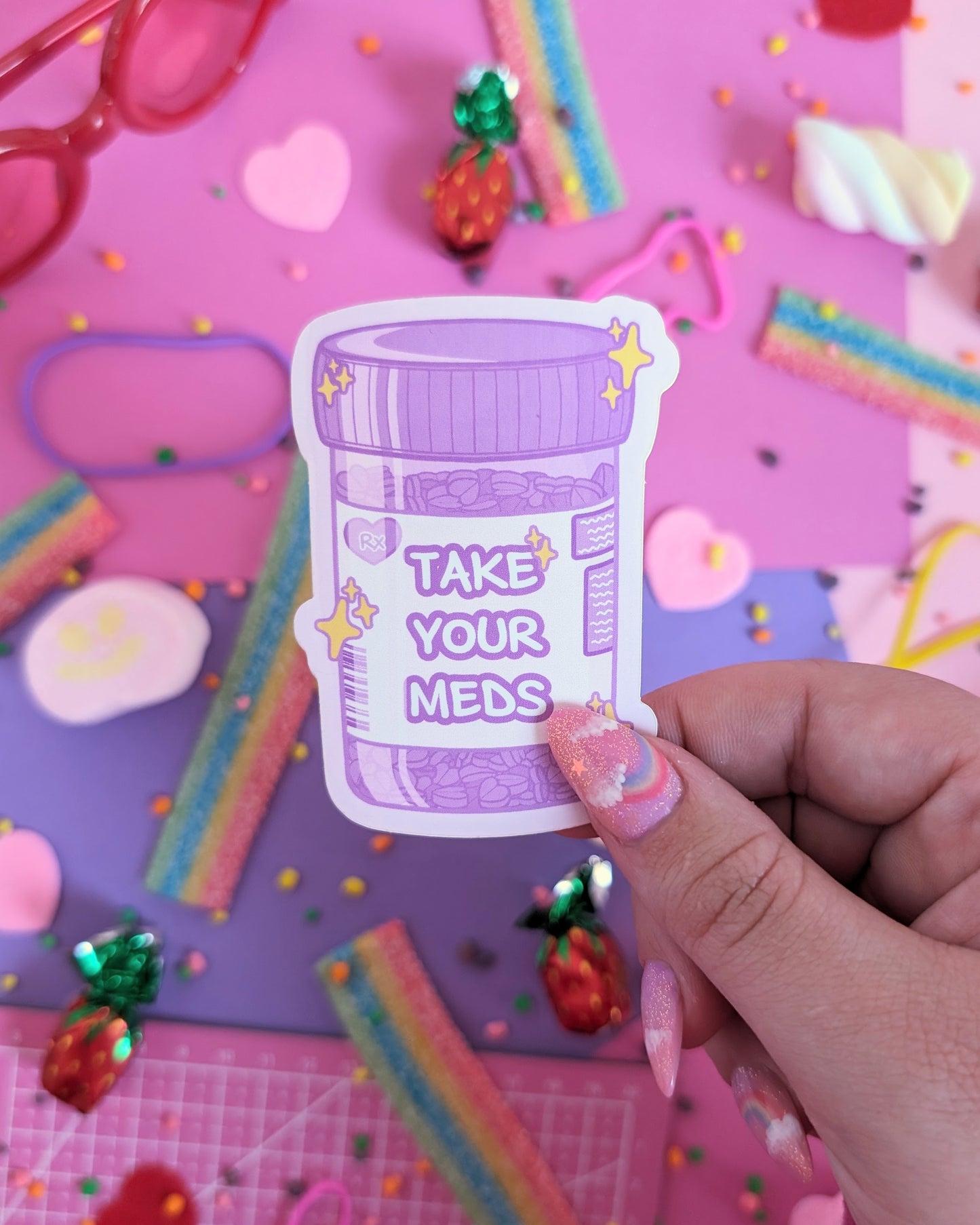 A hand with pink nails holds a purple sticker shaped like a prescription bottle saying "Take Your Meds", set against a pink and purple background with rainbow candies