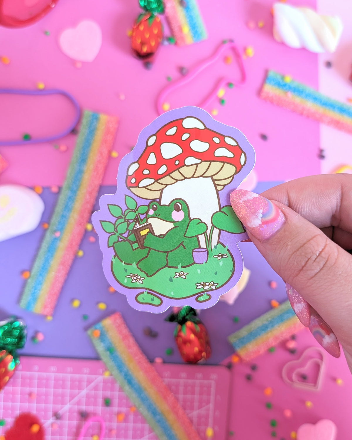 A hand with pink glittery nails holds a sticker featuring a green frog reading a book under a red-and-white mushroom. The backdrop is a colourful pink and purple background with candies.
