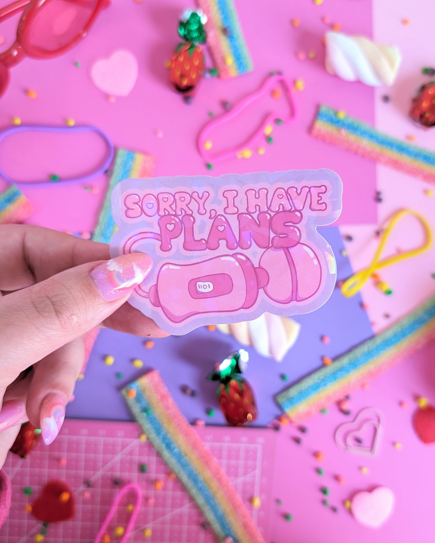 A hand with pink nails holds a sparkly sticker of a vibrator saying, "Sorry, I have plans," set over a pink and purple background with rainbow candies.