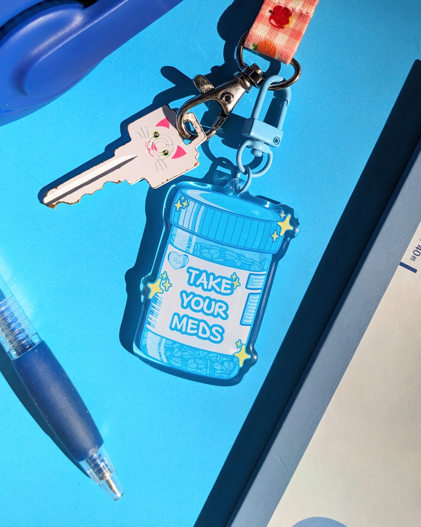 A blue keychain charm shaped like a prescription bottle saying "Take Your Meds" text against a blue background