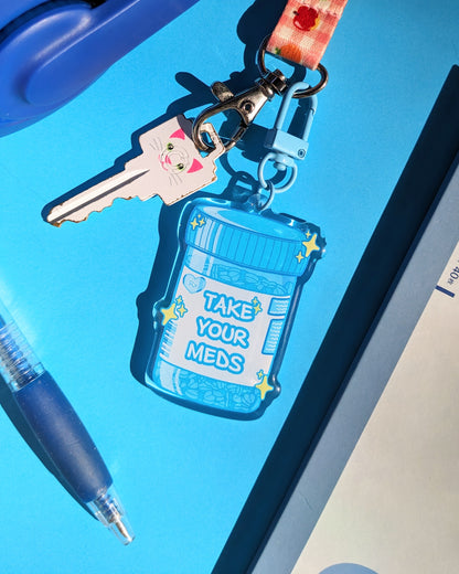 A blue keychain charm shaped like a prescription bottle saying "Take Your Meds" text against a blue background