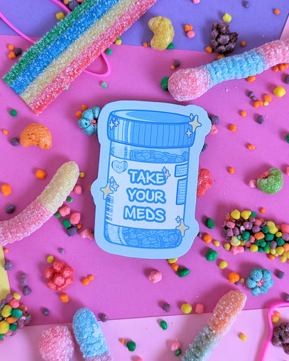 Blue magnet shaped like a prescription bottle that says "Take Your Meds" set against a pink and purple background with rainbow candies