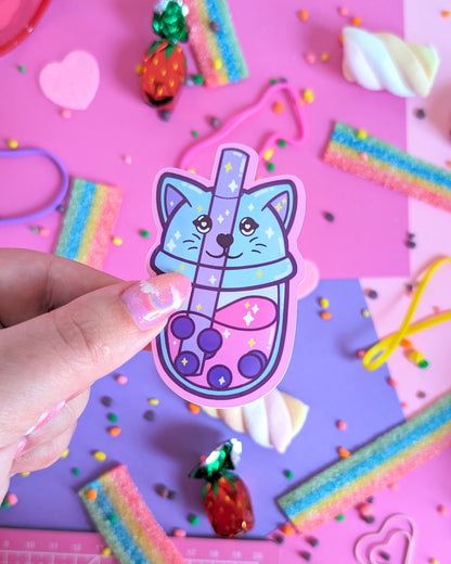 A hand with glittery pink nails holds a  sticker featuring a stylized bubble tea with a lid shapes like a cat head. The backdrop is pink and purple with colourful candies.