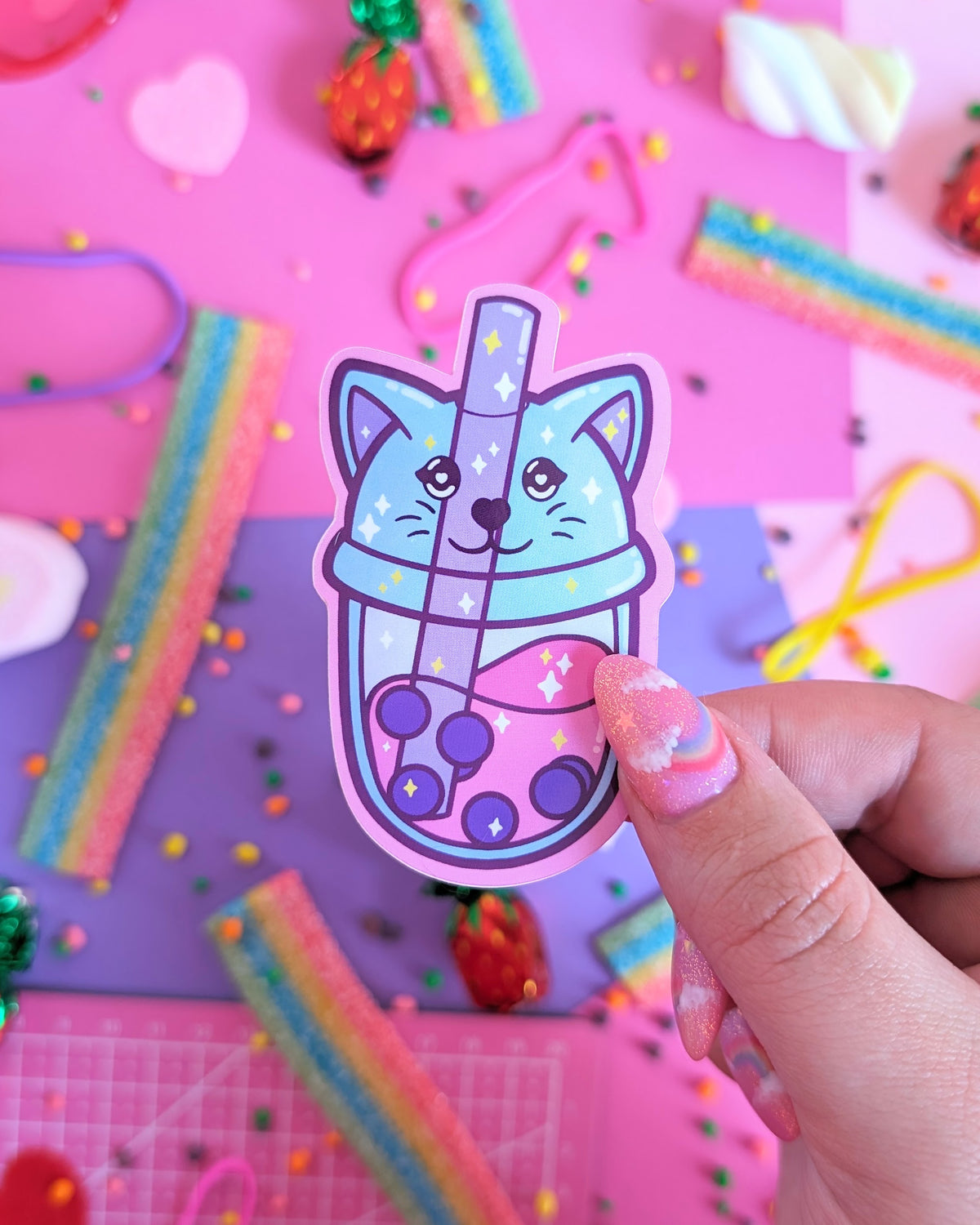 A hand with glittery pink nails holds a  sticker featuring a stylized bubble tea with a lid shapes like a cat head. The backdrop is pink and purple with colourful candies.