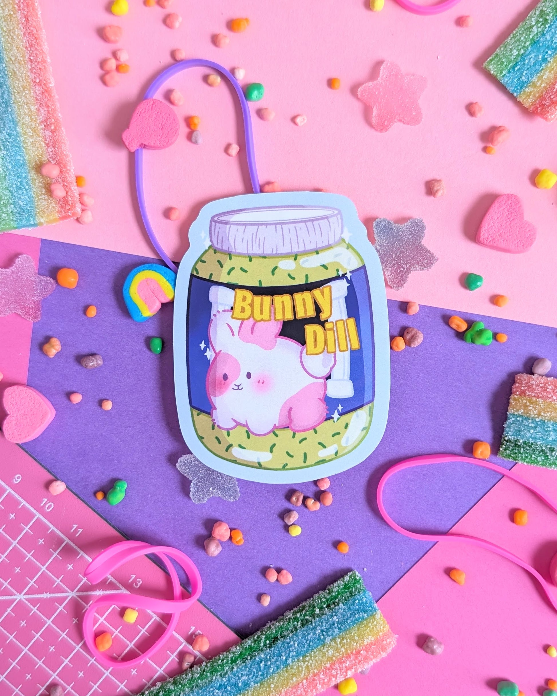 Sticker of a cute pink bunny in a sauce jar labeled "Bunny Dill" on pastel pink and purple background. Surrounded by colourful candy.