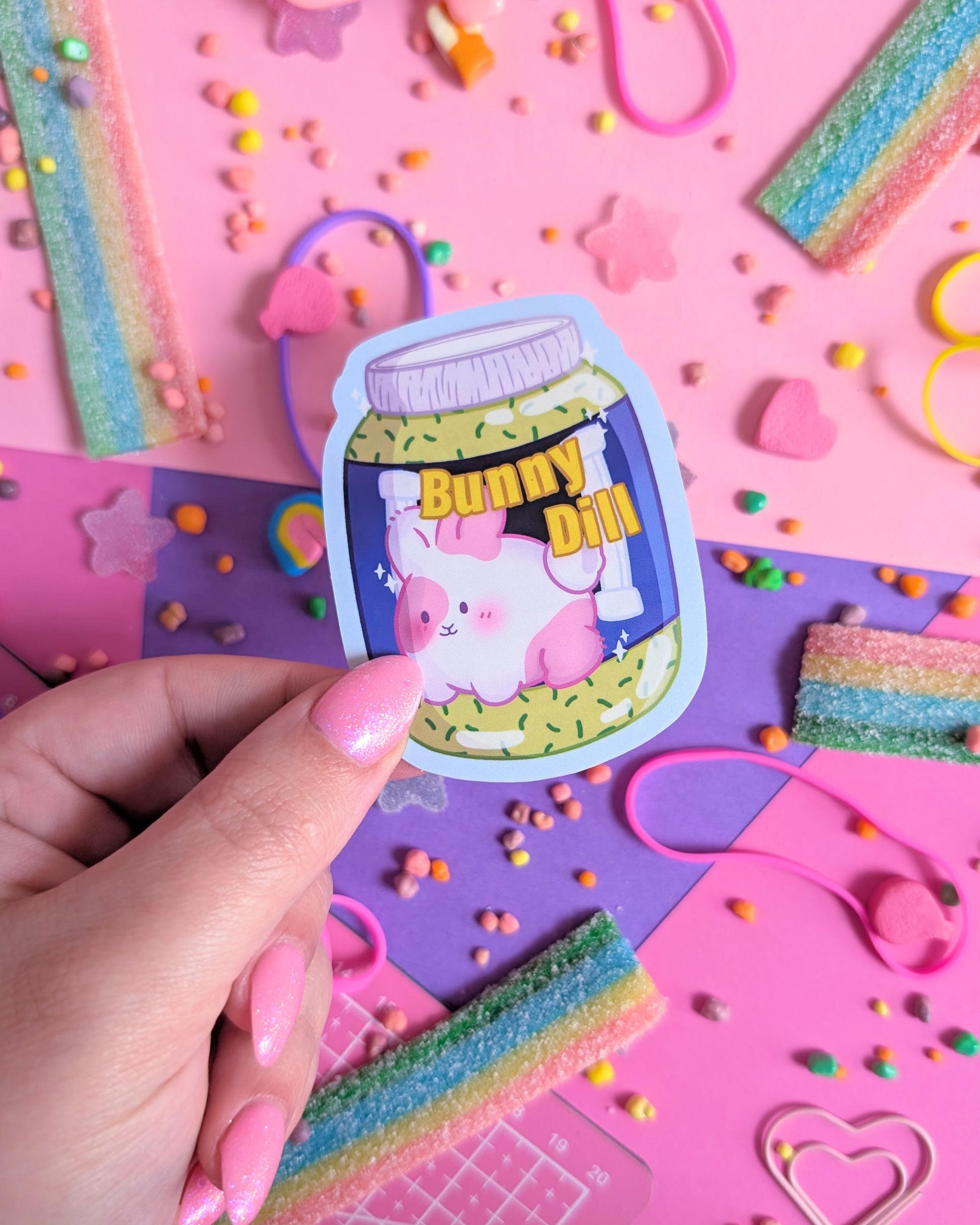 Hand with pink nails hold a sticker of a cute pink bunny in a sauce jar labeled "Bunny Dill" on pastel pink and purple background. Surrounded by colourful candy.