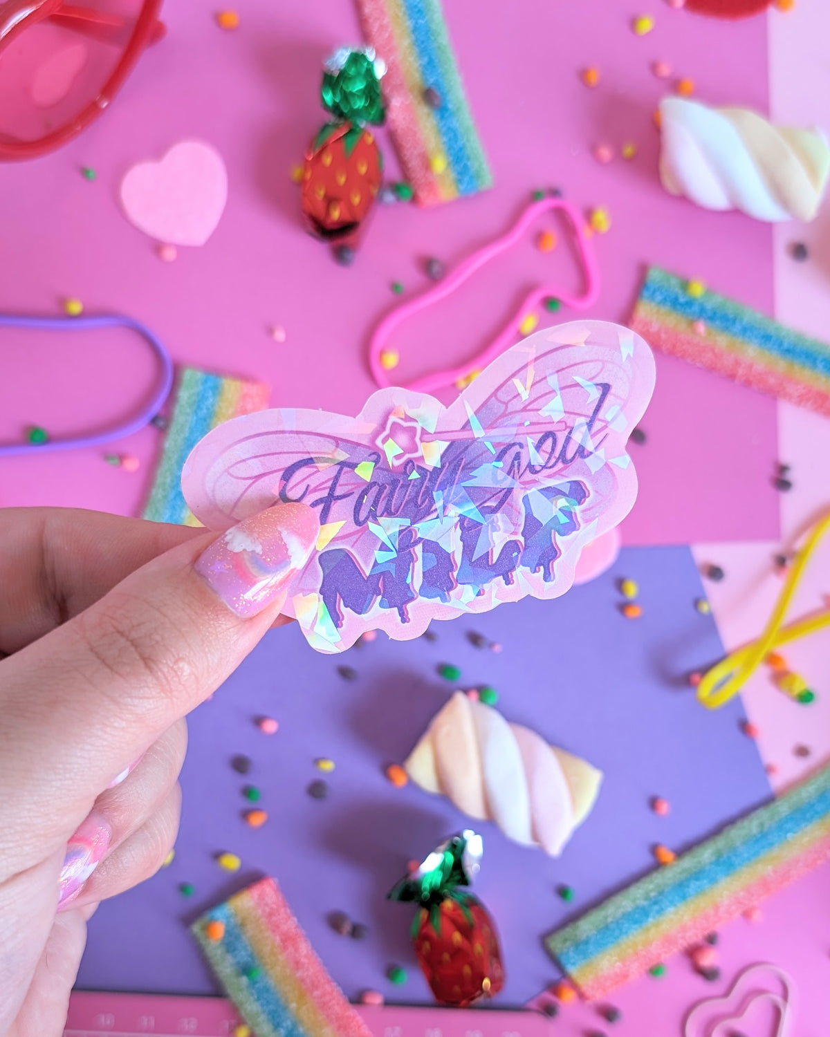 A hand with glittery pink nails holds a holographic butterfly-shaped sticker with the text 'Fairy god MILK' in purple glittery lettering with a magic wand on top. The background is a colourful flatlay of pink and purple sheets decorated with candies.