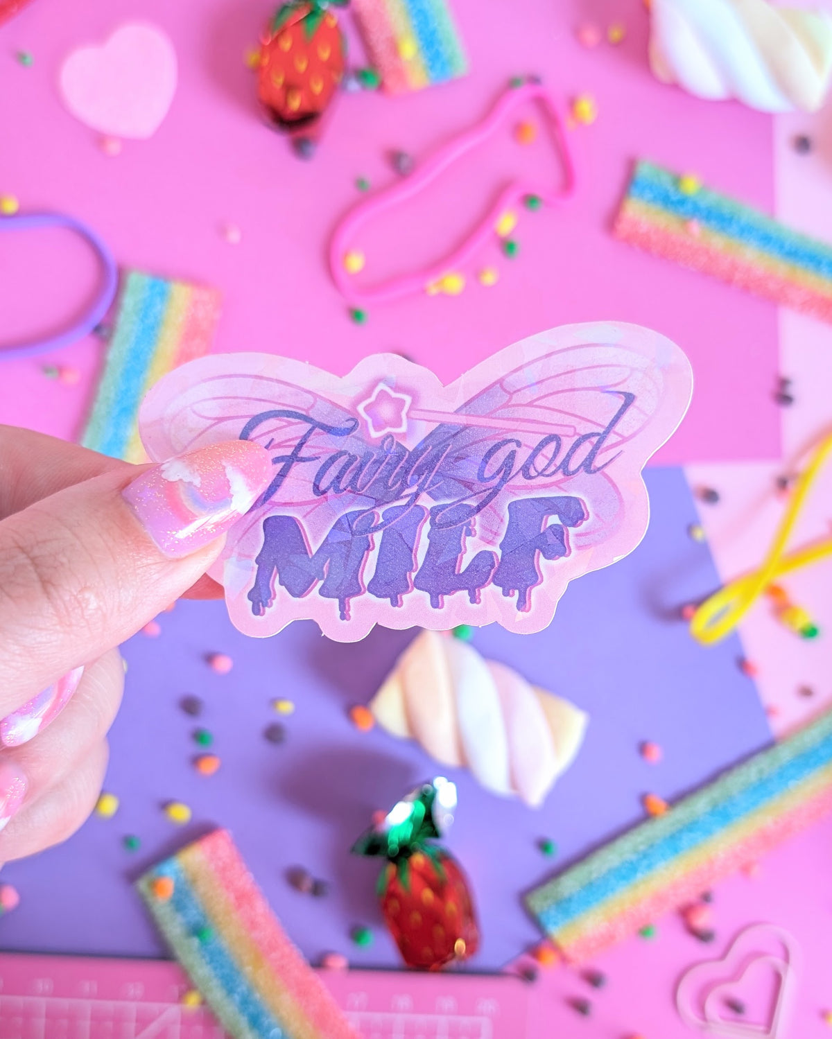 A hand with glittery pink nails holds a holographic butterfly-shaped sticker with the text 'Fairy god MILK' in purple glittery lettering with a magic wand on top. The background is a colourful flatlay of pink and purple sheets decorated with candies.
