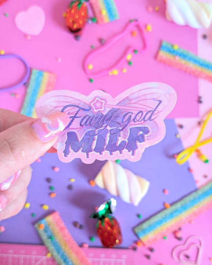 A hand with glittery pink nails holds a holographic butterfly-shaped sticker with the text 'Fairy god MILK' in purple glittery lettering with a magic wand on top. The background is a colourful flatlay of pink and purple sheets decorated with candies.