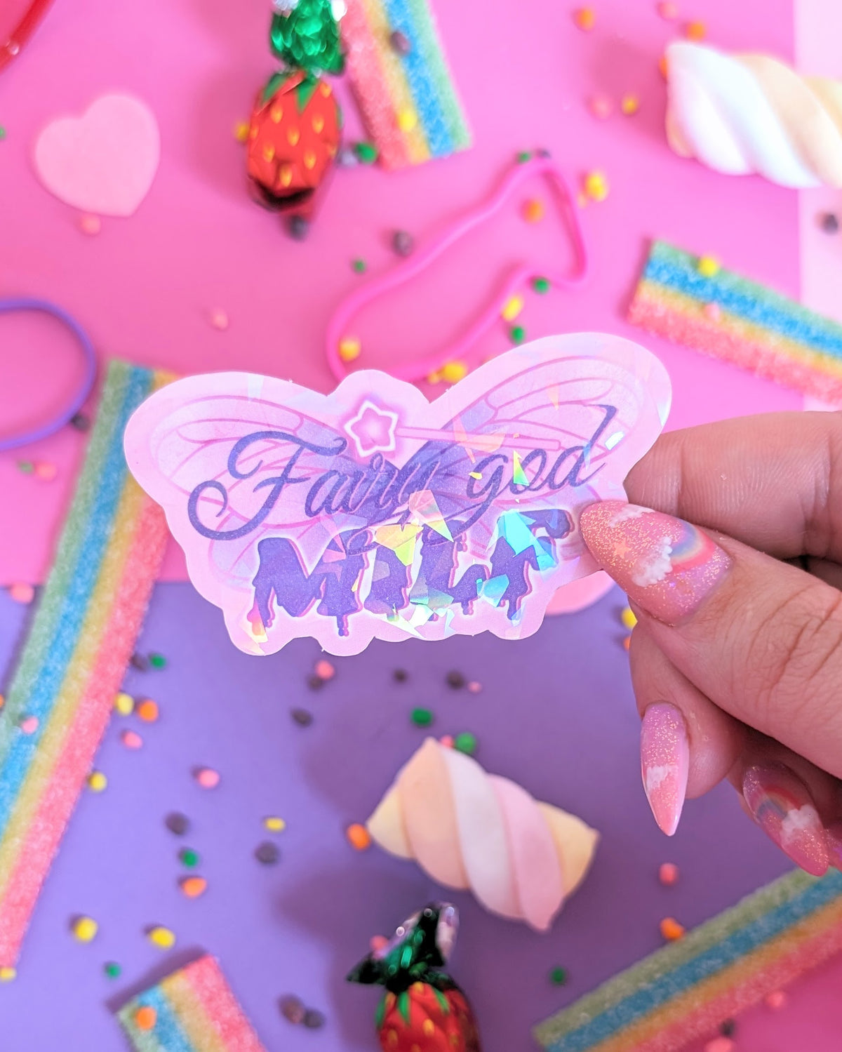 A hand with glittery pink nails holds a holographic butterfly-shaped sticker with the text 'Fairy god MILK' in purple glittery lettering with a magic wand on top. The background is a colourful flatlay of pink and purple sheets decorated with candies.