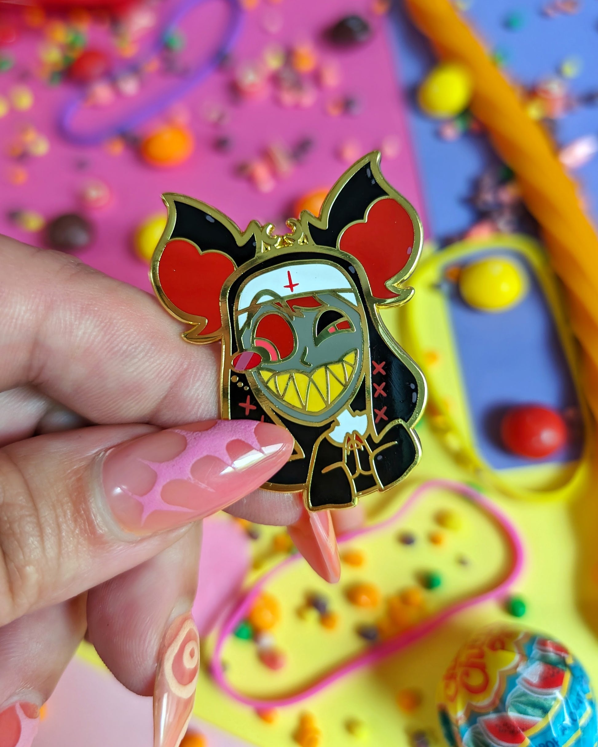 Hand with pink nails holding enamel pin of Alastor from Hazbin Hotel set against a colourful background with rainbow candies