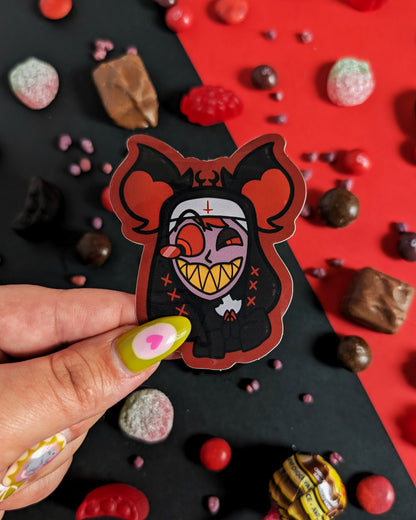 Hand holding a sticker of Alastor from Hazbin Hotel dressed as a nun against red and black background with red, black, and brown candies.