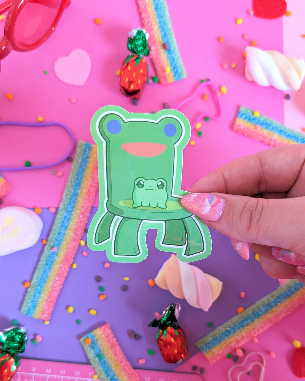 A hand with pink glittery nails holds a green frog-shaped sticker or decal with a smaller frog graphic in the center. The background is a colorful flat-lay styled with candy, rainbow strips, sprinkles, and pastel props on a pink and purple surface.