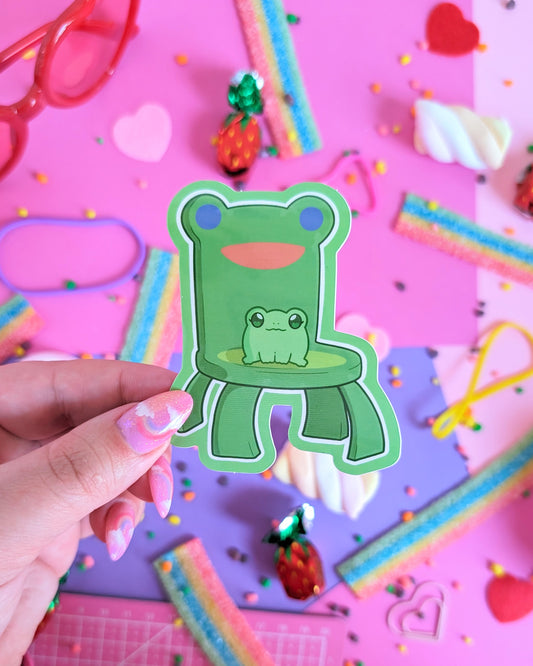 A hand with pink glittery nails holds a green frog-shaped sticker or decal with a smaller frog graphic in the center. The background is a colorful flat-lay styled with candy, rainbow strips, sprinkles, and pastel props on a pink and purple surface.