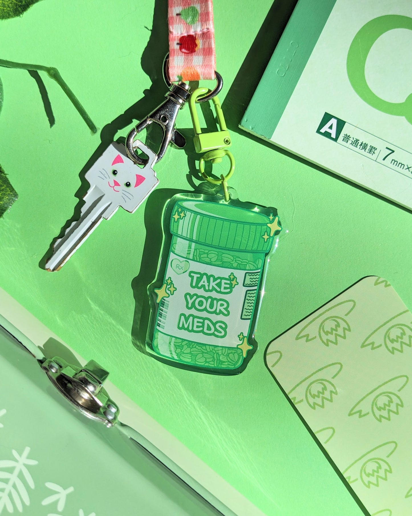 A green keychain charm shaped like a prescription bottle saying "Take Your Meds" text against a green background