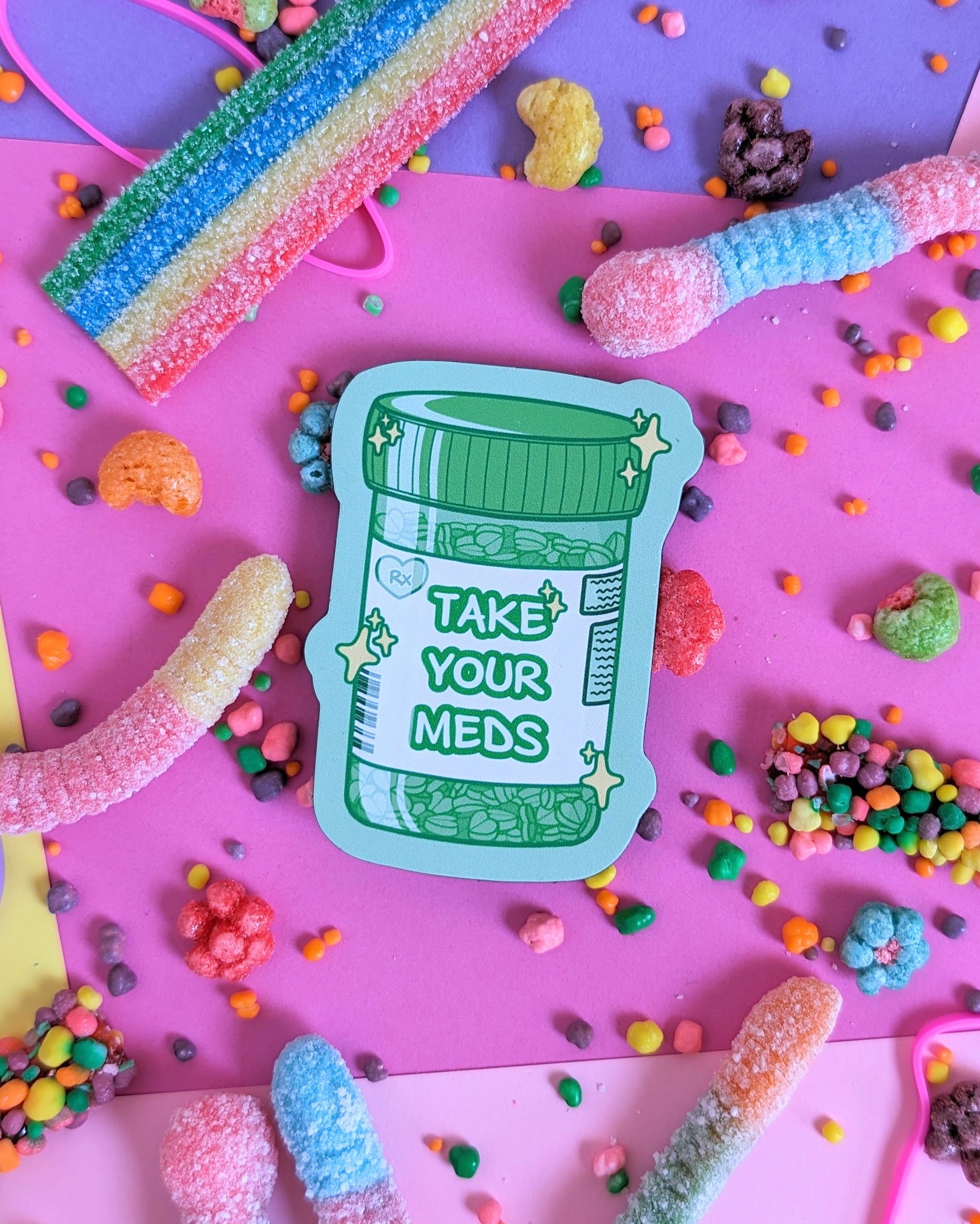 Green magnet shaped like a prescription bottle that says "Take Your Meds" set against a pink and purple background with rainbow candies