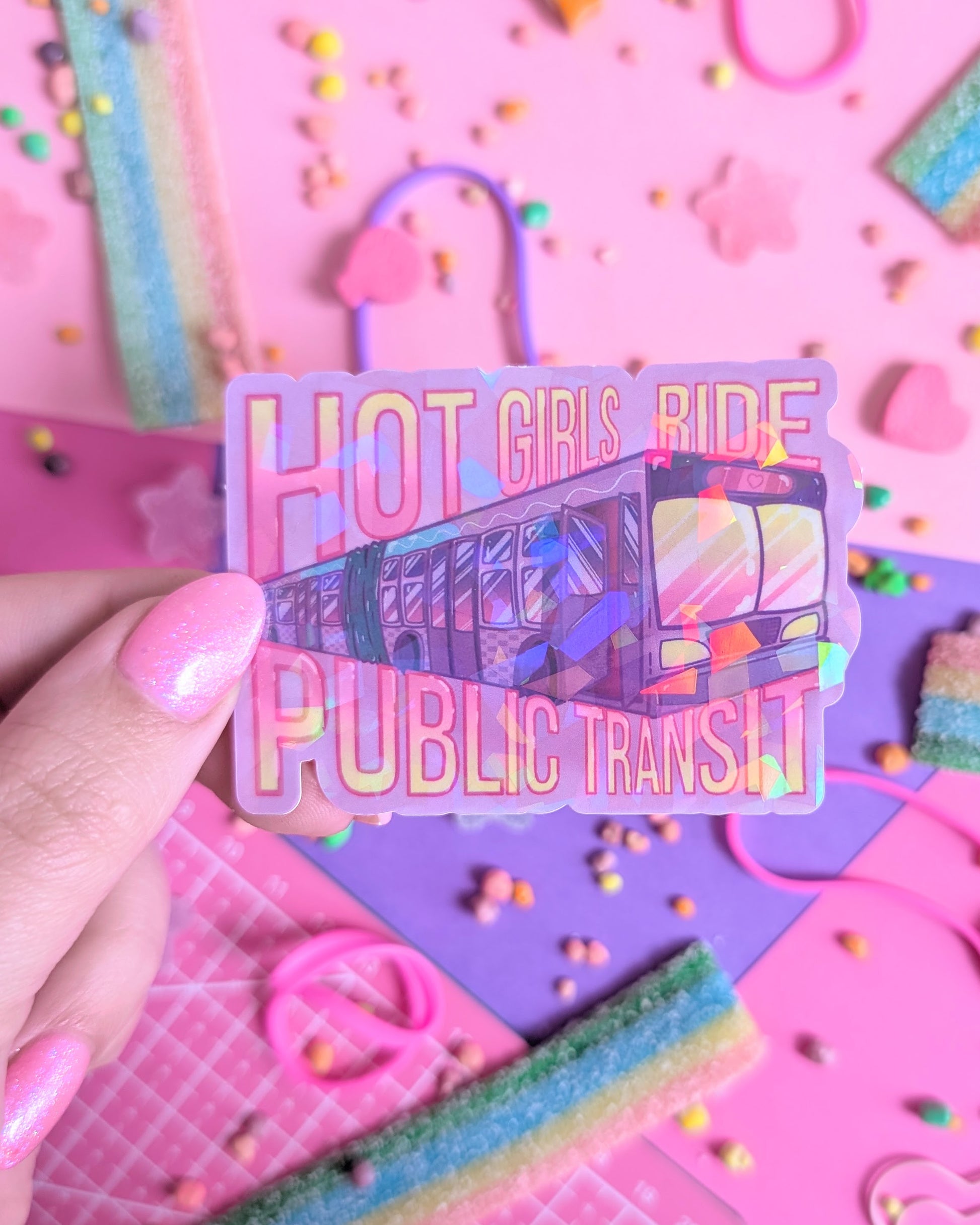 A hand with pink nail polish holds a colourful, sparkly  sticker of a bus with the text "Hot Girls Ride Public Transit." The background is pink and purple with rainbow candies.