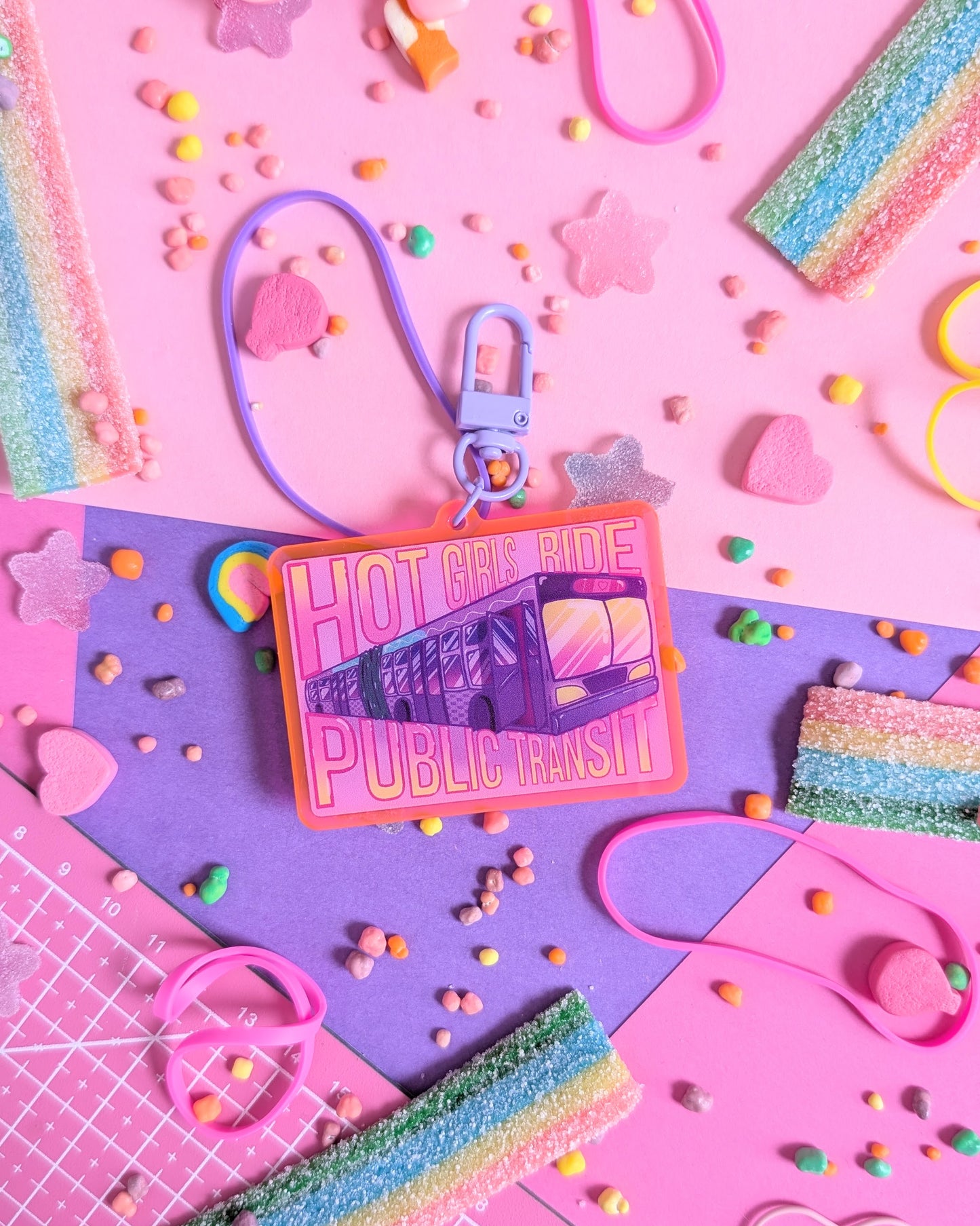 Keychain charm with "Hot Girls Ride Public Transit" text over a transit bus. Background is pink and purple with colourful candies
