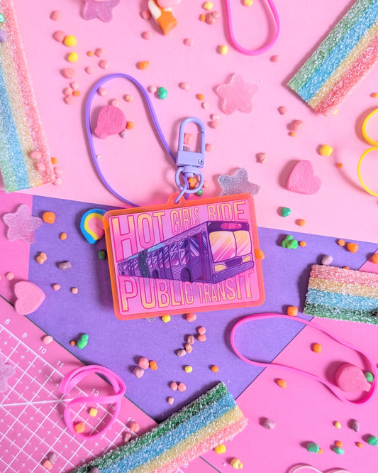 Keychain charm with "Hot Girls Ride Public Transit" text over a transit bus. Background is pink and purple with colourful candies