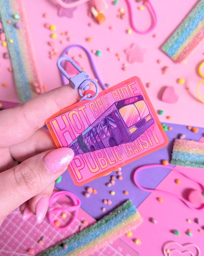 Hand holding a keychain charm with "Hot Girls Ride Public Transit" text over a transit bus. Background is pink and purple with colourful candies