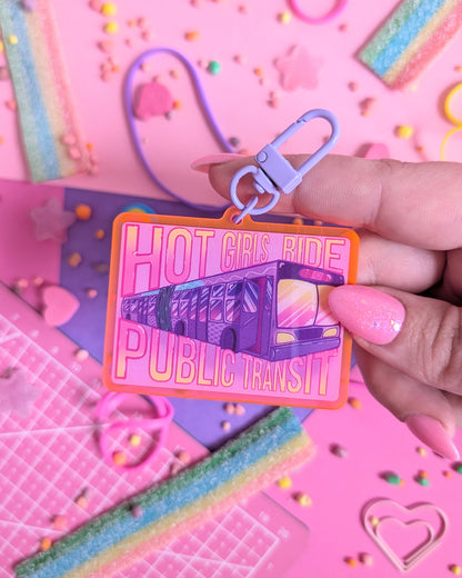 Hand holding a keychain charm with "Hot Girls Ride Public Transit" text over a transit bus. Background is pink and purple with colourful candies