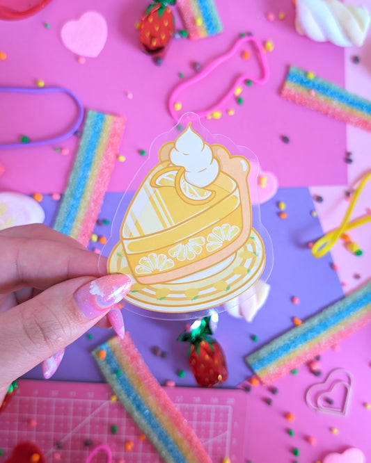 A hand with glittery pink nails holds a sticker featuring a cartoon lemon pie. The backdrop is pink and purple with colourful candies.