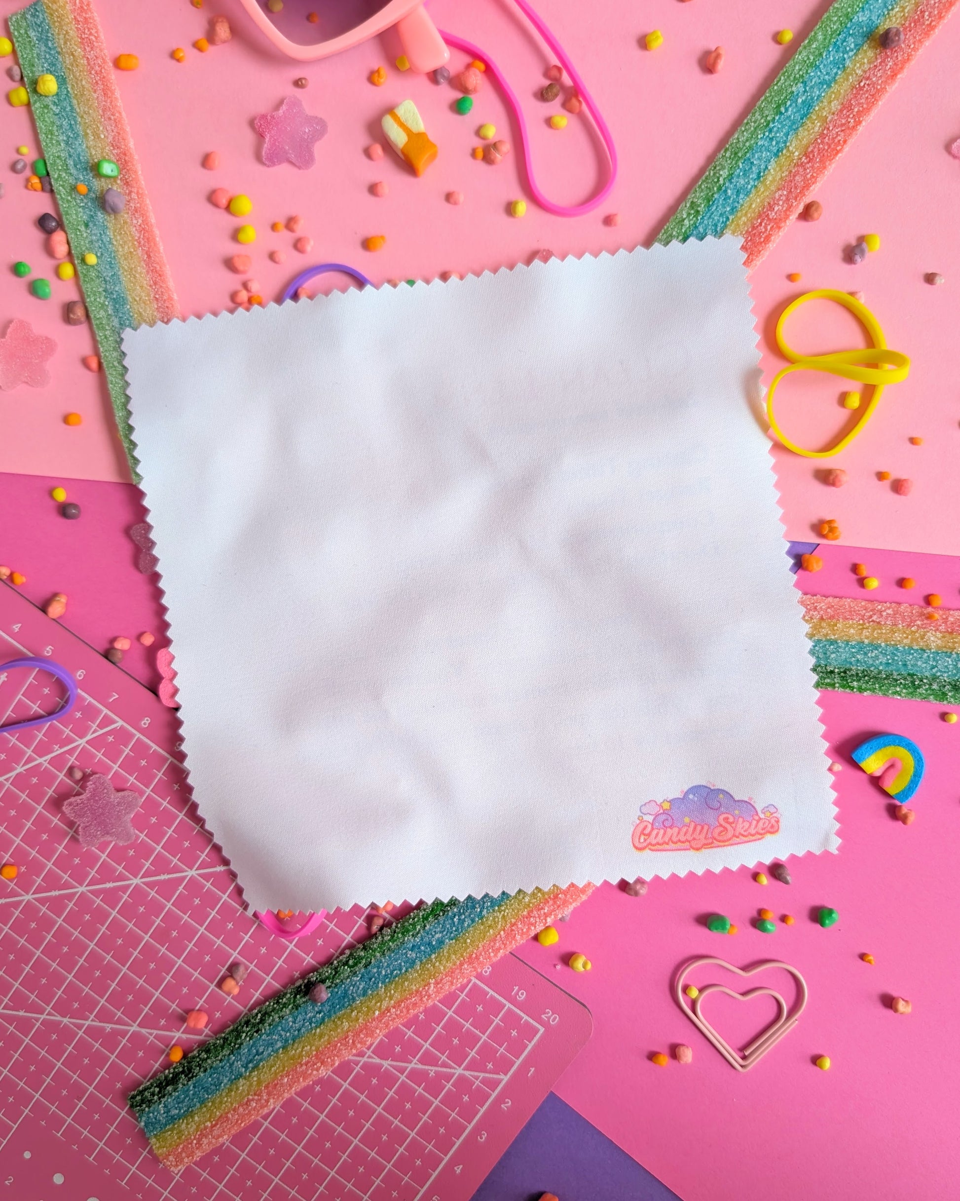 Plain white backside of a lens wipe cloth with the Candy Skies logo on a pink background filled with colourful candies