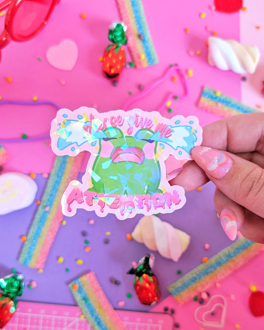 Hand with pink nails holding a colorful sticker with a green cartoon frog and the text "please give me ATTENTION" on a vibrant pink and purple candy-themed background.