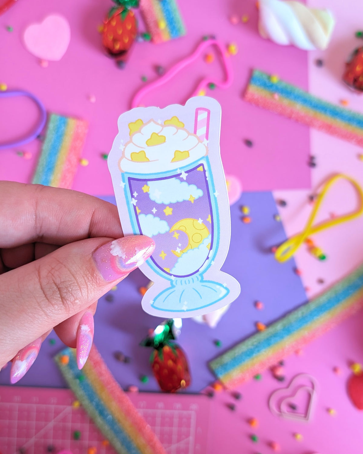 A hand with glittery pink nails holds a sticker of a milkshake containing clouds and stars. The background is a colourful flatlay of pink and purple sheets decorated with candies.