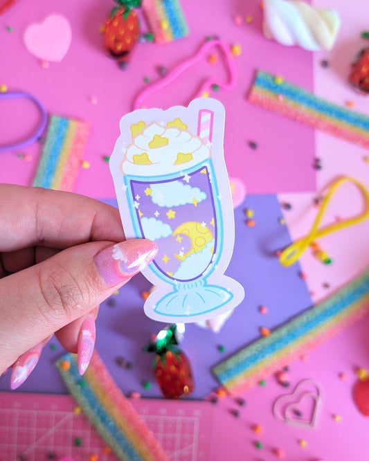 A hand with glittery pink nails holds a sticker of a milkshake containing clouds and stars. The background is a colourful flatlay of pink and purple sheets decorated with candies.
