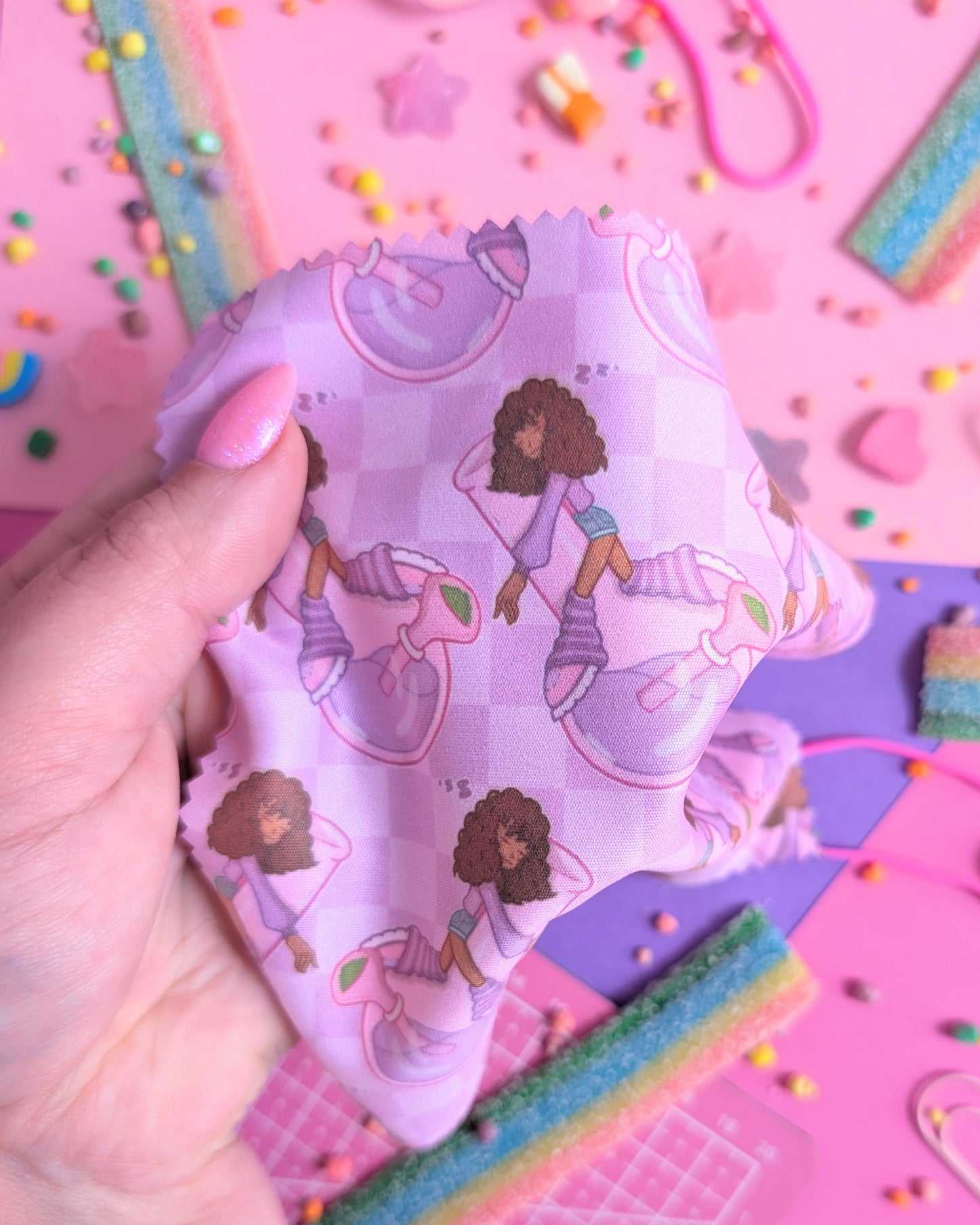 Hand with pink nails holding a draped purple lens wipe cloth with a miniature girl sleeping on a bubbler pattern set against a pink and purple background filled with rainbow candies