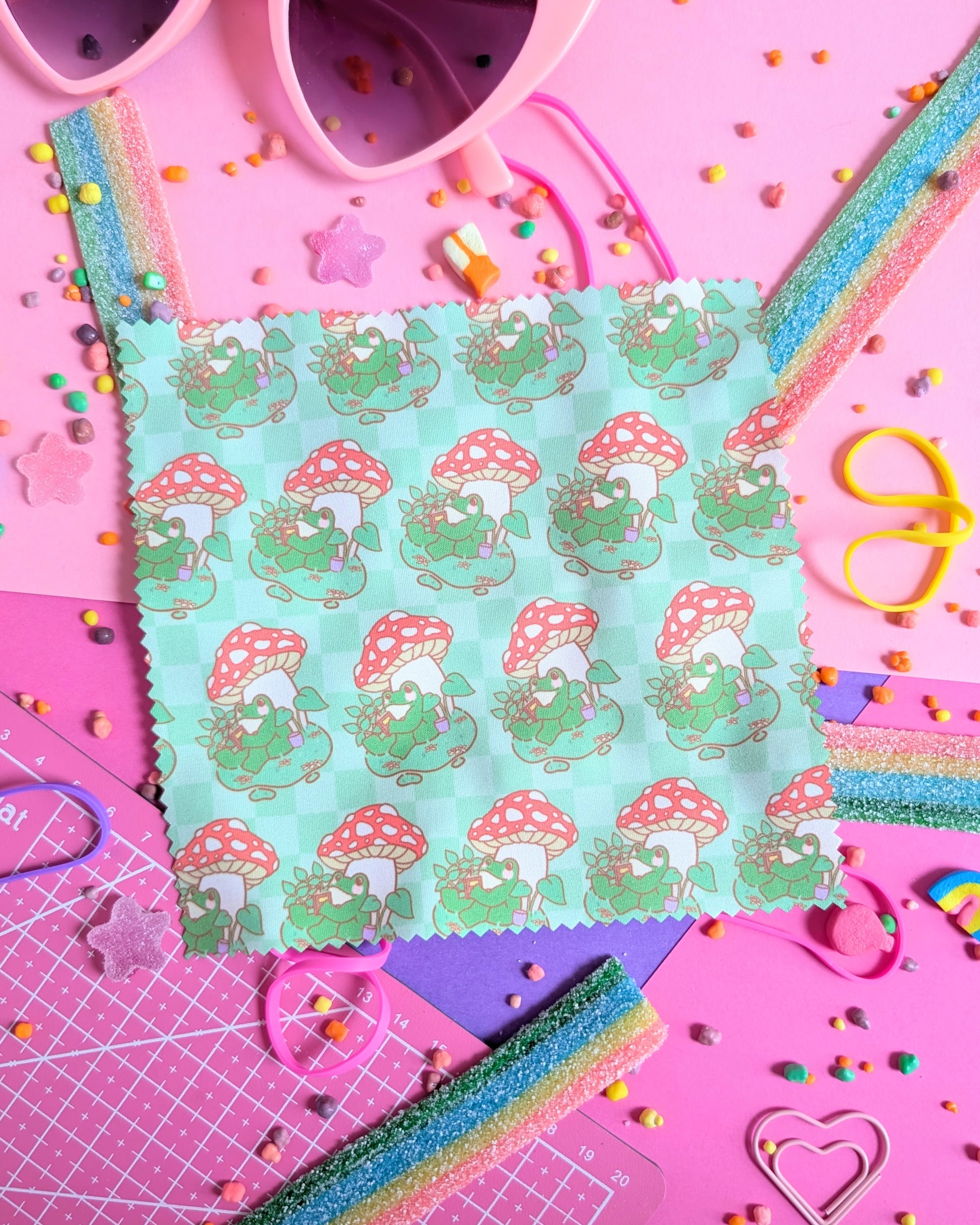 Green lens wipe cloth with a pattern of a frog under a mushroom reading a book set against a pink and purple background filled with rainbow candies