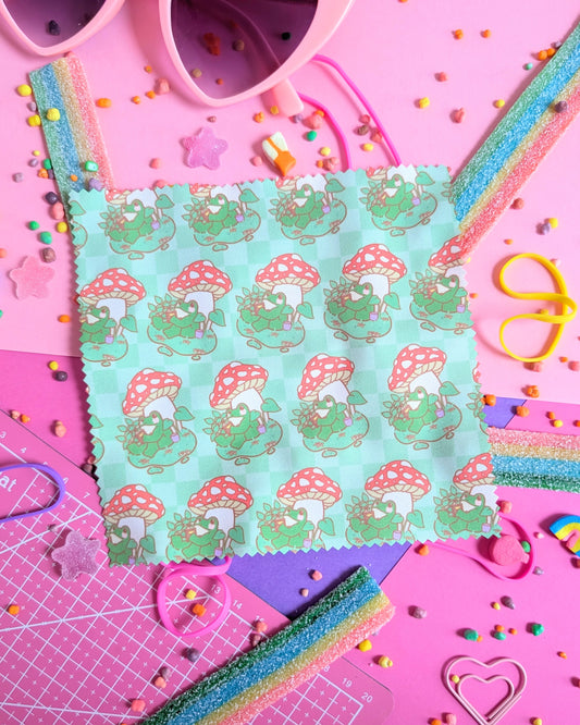 Green lens wipe cloth with a pattern of a frog under a mushroom reading a book set against a pink and purple background filled with rainbow candies