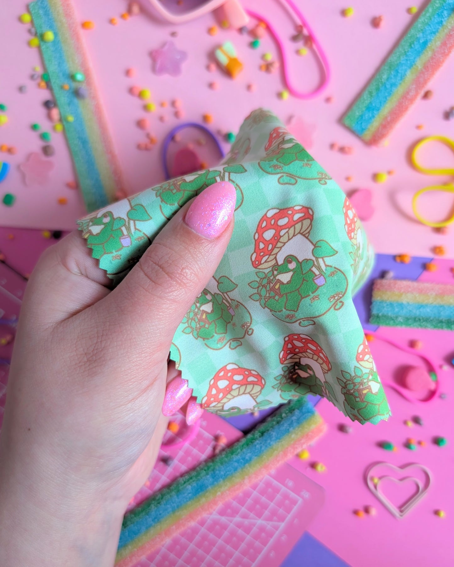 Hand with pink nails holding a draped green lens wipe cloth with a pattern of a frog under a mushroom reading a book set against a pink and purple background filled with rainbow candies