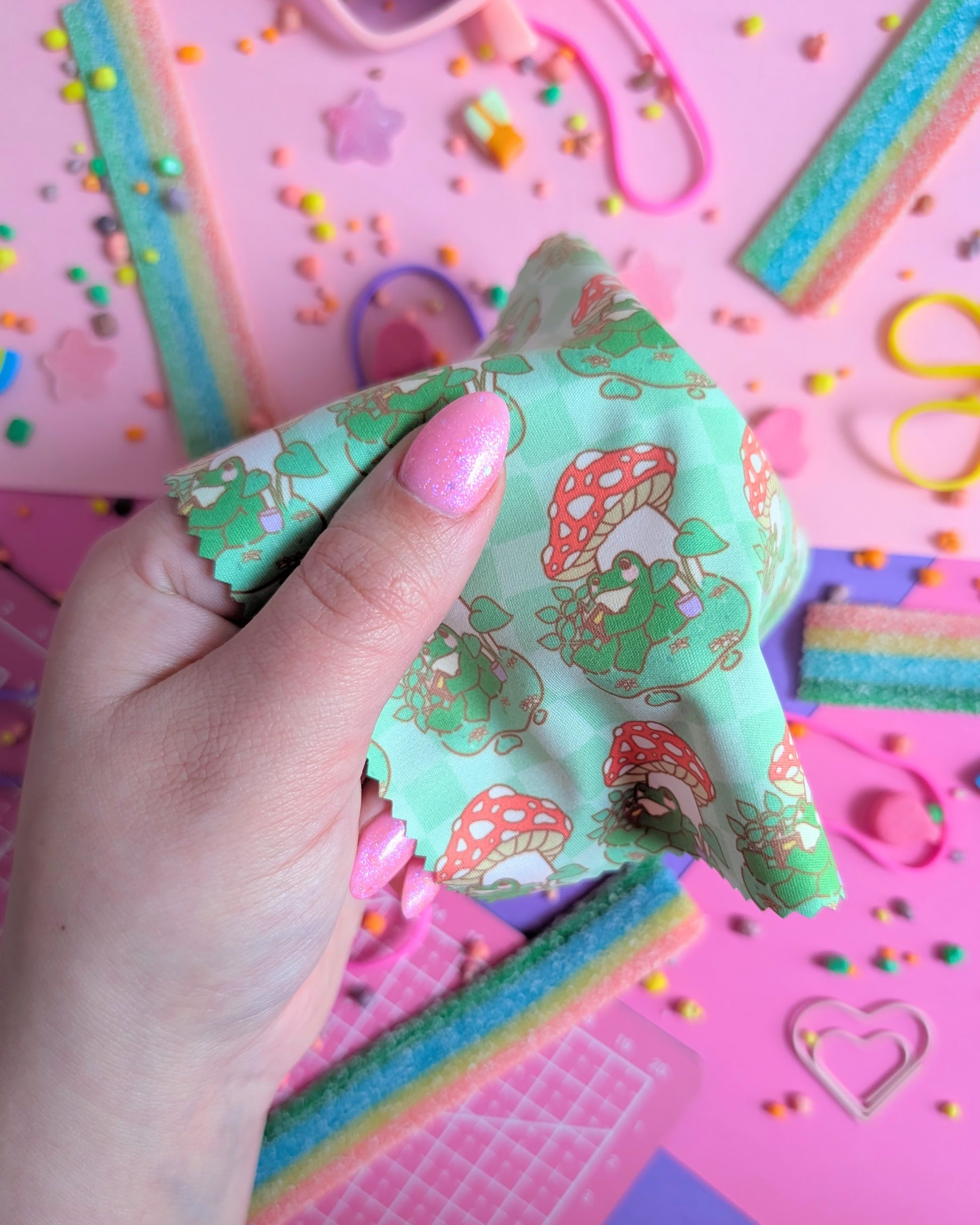 Hand with pink nails holding a draped green lens wipe cloth with a pattern of a frog under a mushroom reading a book set against a pink and purple background filled with rainbow candies