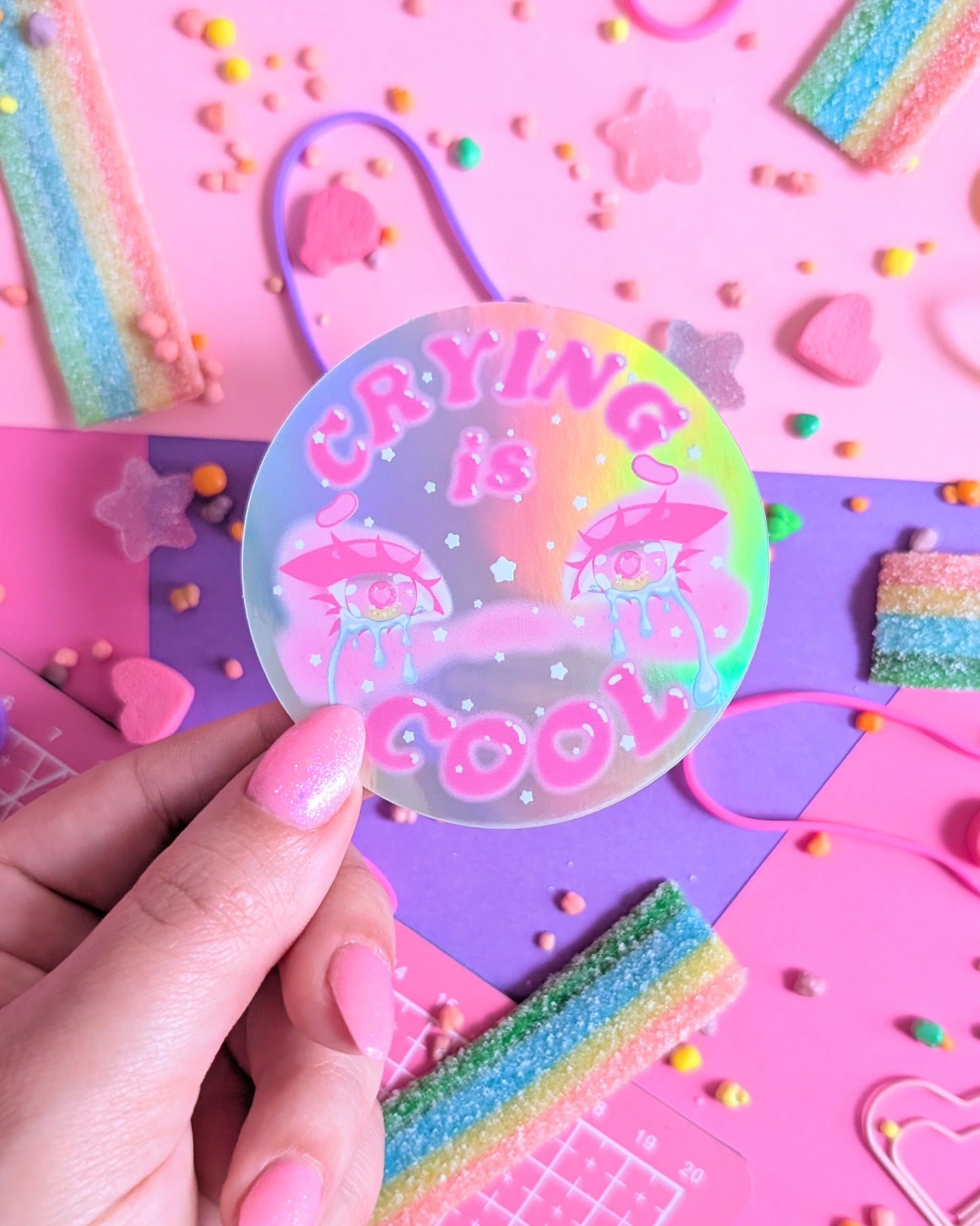 A hand with baby pink nails hold a colourful round sticker with "Crying is Cool" text in pink and two cartoon eyes with tears on a neon rainbow background. Surrounded by pastel candy on pink and purple paper.