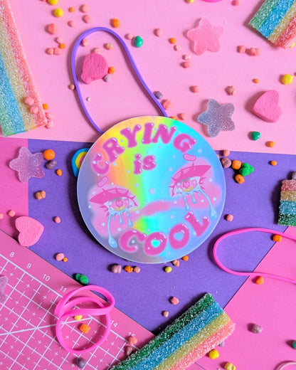 A colorful round sticker with "Crying is Cool" text in pink and two cartoon eyes with tears on a neon rainbow background. Surrounded by pastel candy on pink and purple paper.