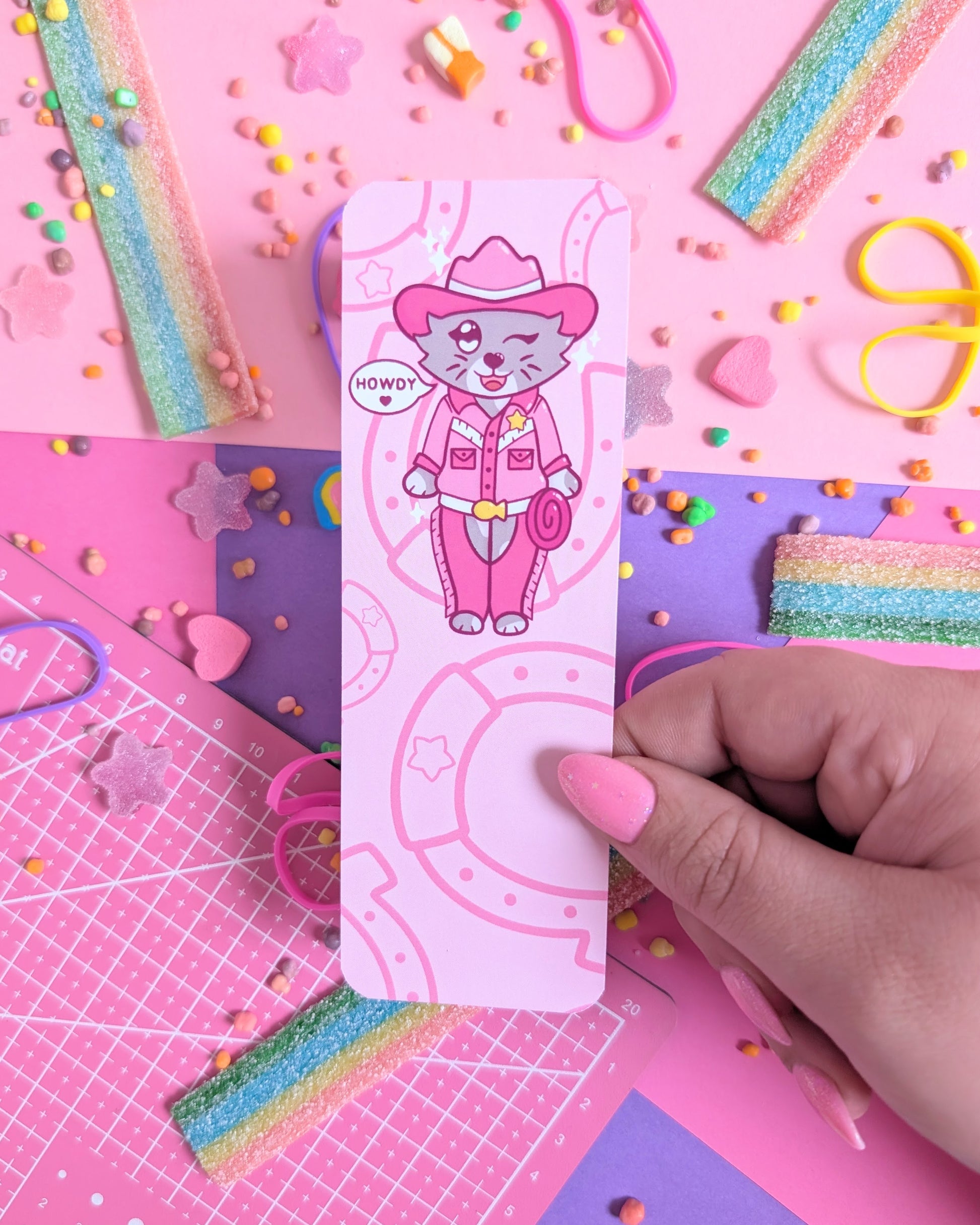 Hand with pink nails holdin a bookmark with a cartoon cat in pink cowboy attire, surrounded by a pink and purple background of colourful candy