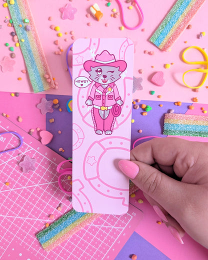 Hand with pink nails holdin a bookmark with a cartoon cat in pink cowboy attire, surrounded by a pink and purple background of colourful candy