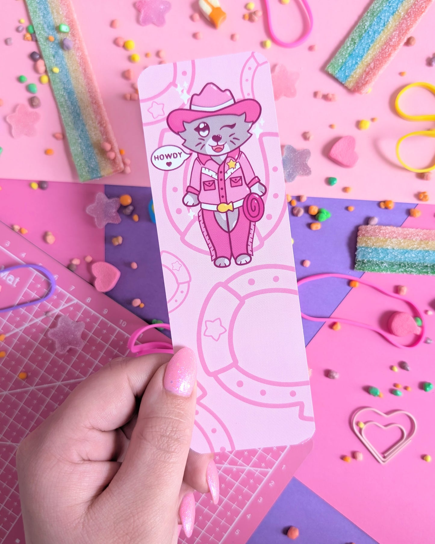 Hand with pink nails holding a bookmark with a cartoon cat in pink cowboy attire, surrounded by a pink and purple background of colourful candy