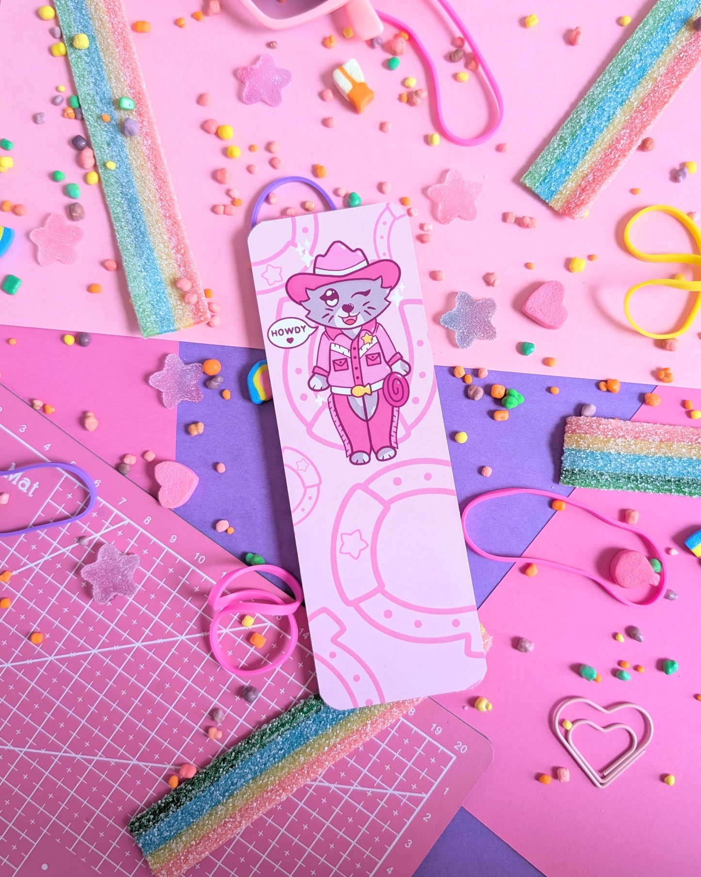 Bookmark with a cartoon cat in pink cowboy attire, surrounded by a pink and purple background of colourful candy