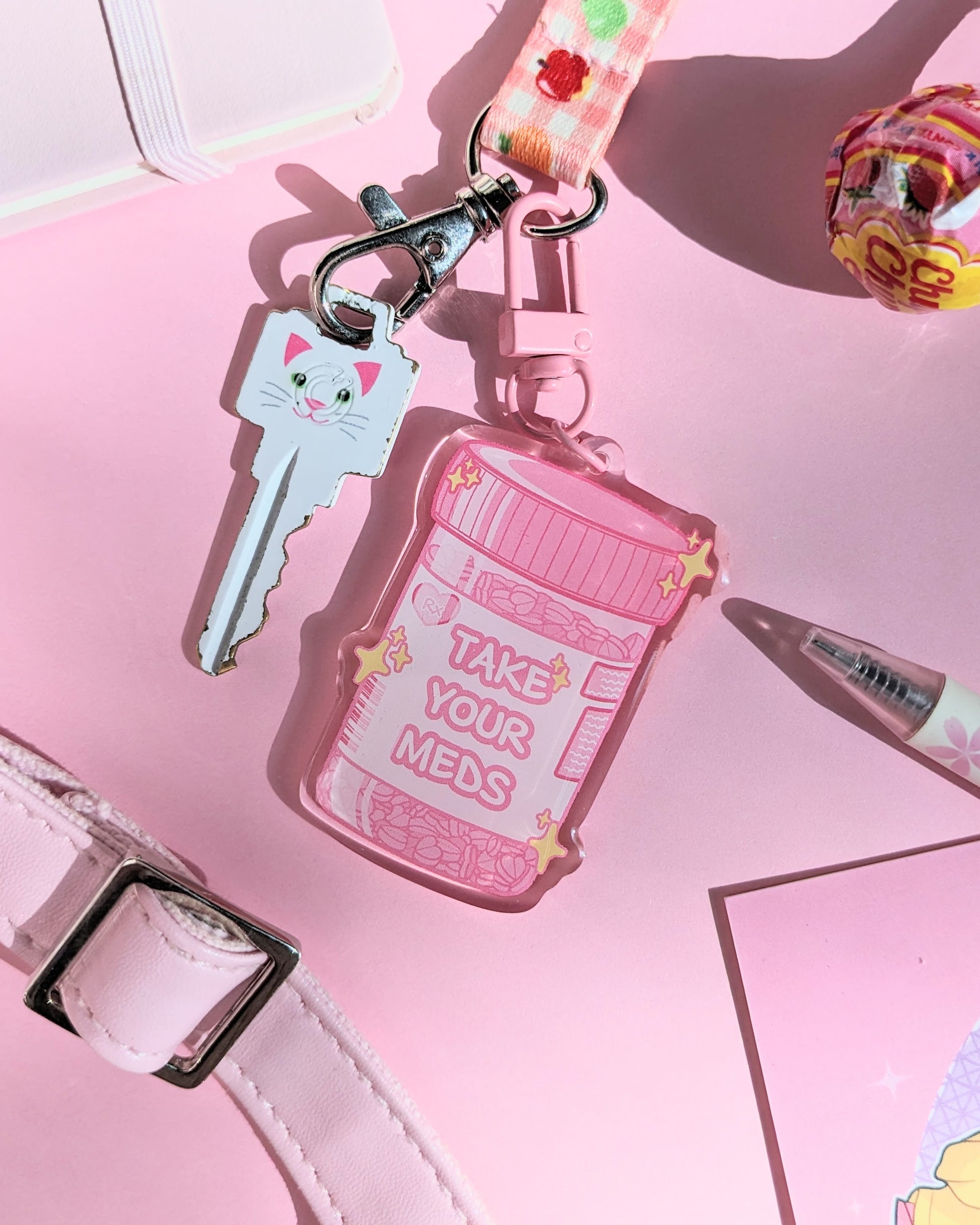 A pink keychain charm shaped like a prescription bottle saying "Take Your Meds" text against a pink background