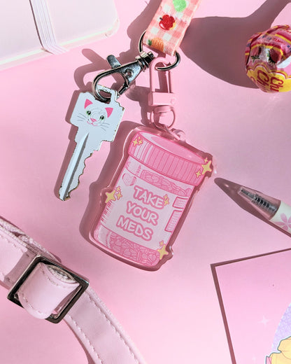 A pink keychain charm shaped like a prescription bottle saying "Take Your Meds" text against a pink background