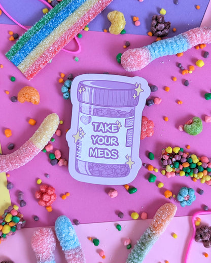 Purple magnet shaped like a prescription bottle that says "Take Your Meds" set against a pink and purple background with rainbow candies