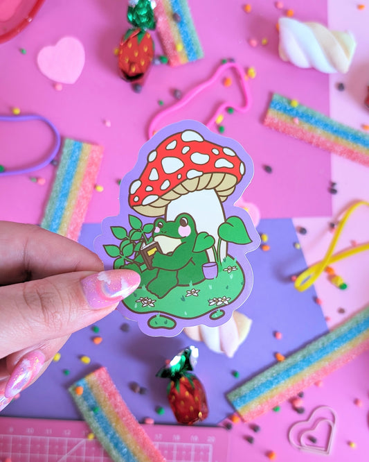 A hand with pink glittery nails holds a sticker featuring a green frog reading a book under a red-and-white mushroom. The backdrop is a colourful pink and purple background with candies.
