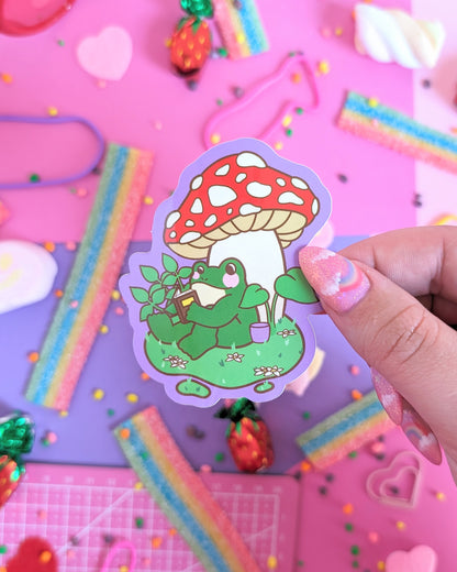 A hand with pink glittery nails holds a sticker featuring a green frog reading a book under a red-and-white mushroom. The backdrop is a colourful pink and purple background with candies.